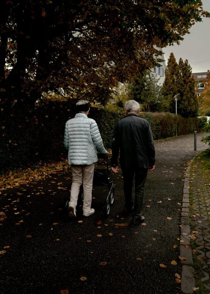 An elderly couple walking together with a walker on a leaf-strewn path in a Munich park, showcasing companionship in autumn.